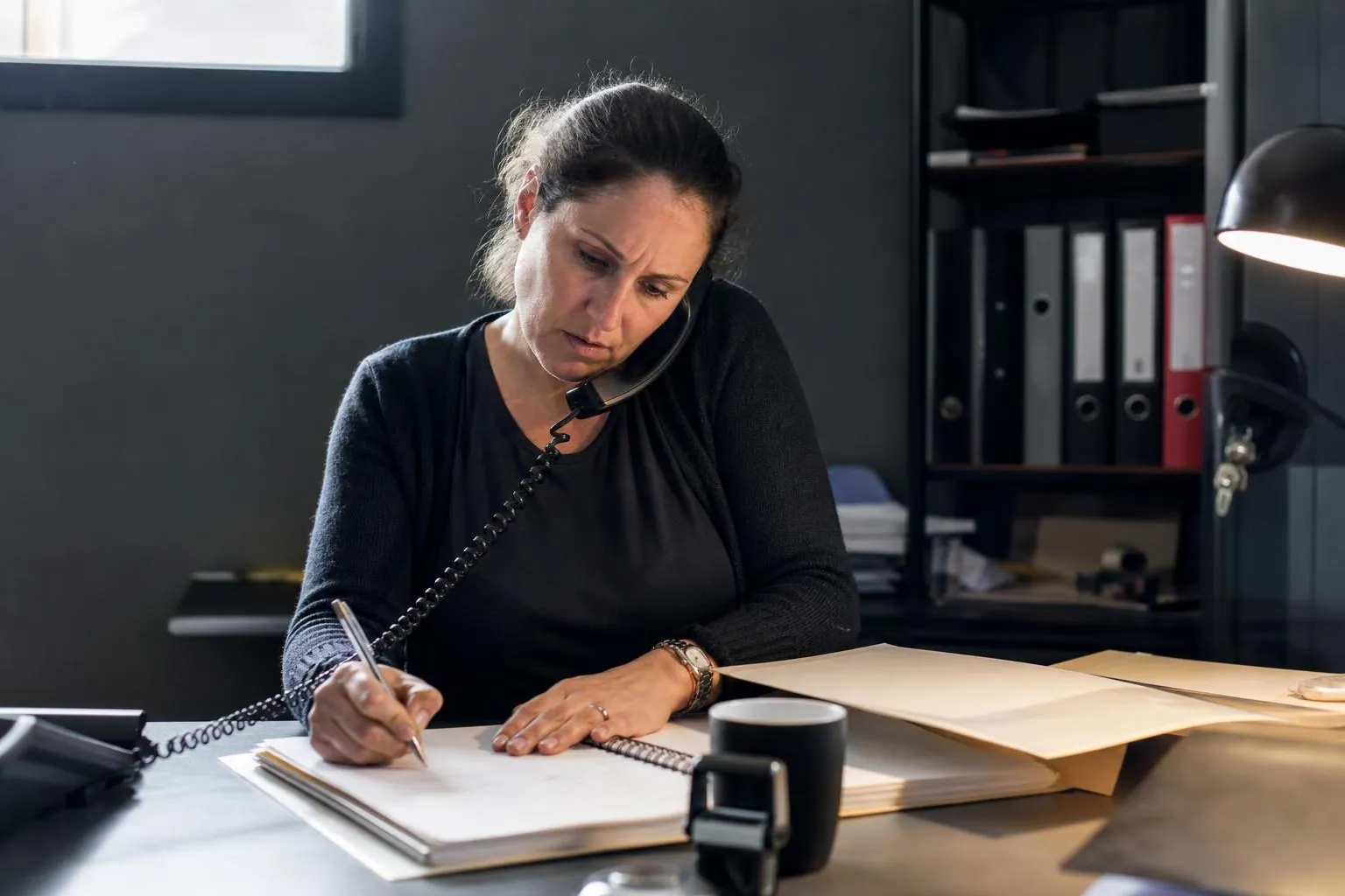Gerante de salle de sport au telephone fixe prenant des notes sur un cahier a spirale dans son bureau